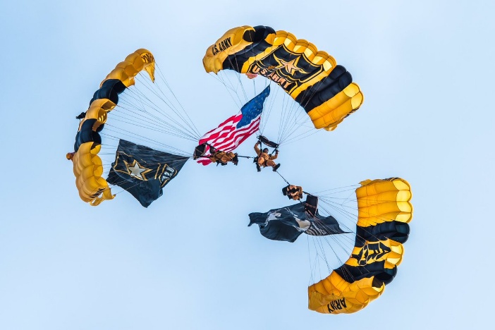 US Army Golden Knights 3 blue and yellow parachutes with US Army logo, three people in yellow jump suits with 1 United States of America flag, 1 US Army flag, all in the sky with a blue background